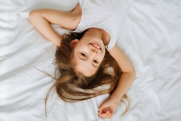 klein ondeugend vrolijk meisje schoolmeisje in de ochtend in een heldere kamer op een witte achtergrond slaapkamer grimaces glimlacht over en gezichten - Foto, afbeelding