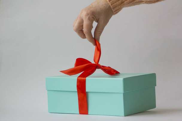 De hand van een oudere vrouw maakt het rode lint van een groene geschenkdoos los op een witte achtergrond - Foto, afbeelding
