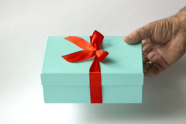 The hand of an elderly man holds out a gift in a light green box tied with a red ribbon on a white background - Fotó, kép