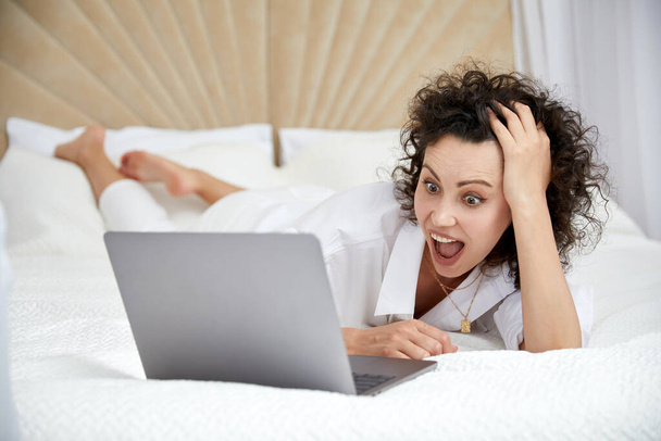 Curly girl using laptop computer sitting on white bed browsing or chatting with friends online - Foto, imagen
