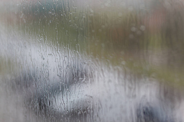 Deszczowy dzień przez okno na zachmurzonym szarym niebie i tle budynków miejskich. Koncepcja. Wieczorny pejzaż za szklanym oknem z miażdżącymi kroplami wody. - Zdjęcie, obraz