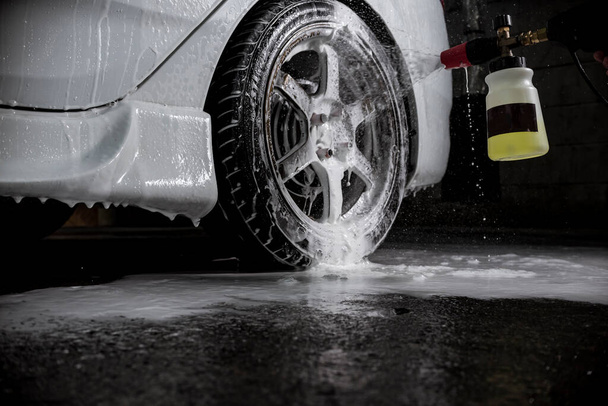 Un hombre utiliza un pulverizador de espuma de coche bomba de mano en las llantas de un coche. En una tienda de lavado de autos o detalles de automóviles. Flash iluminado. - Foto, imagen