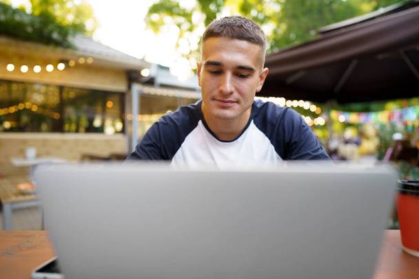 Young man sitting at table and typing on laptop keyboard while working in outdoor cafe - Foto, Imagen