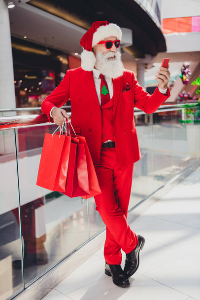 Вертикальный полный размер тела вид на привлекательный бородатый веселый Санта покупает подарки с помощью приложения гаджет заказ в торговом центре в помещении - Фото, изображение
