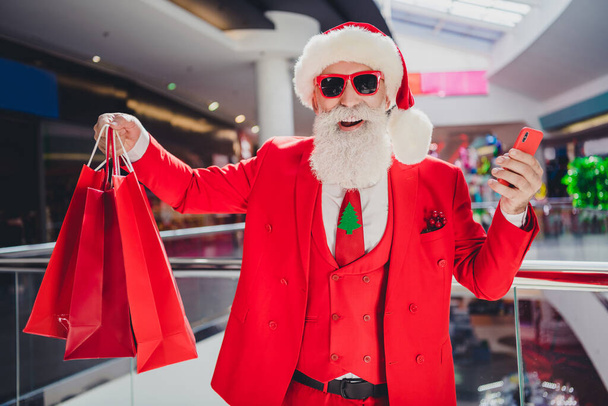 Photo of popular santa hold phone prepare x-mas presents wear sunglass hat red tux in supermarket mall center - Фото, изображение