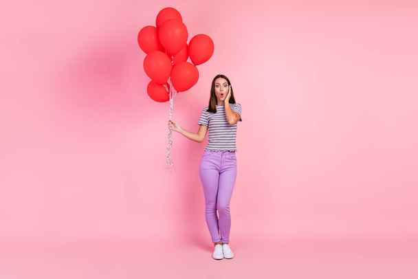 Foto van aantrekkelijke onder de indruk jonge vrouw gekleed gestreepte kleding met veel rode ballonnen arm wang geïsoleerde roze kleur achtergrond - Foto, afbeelding