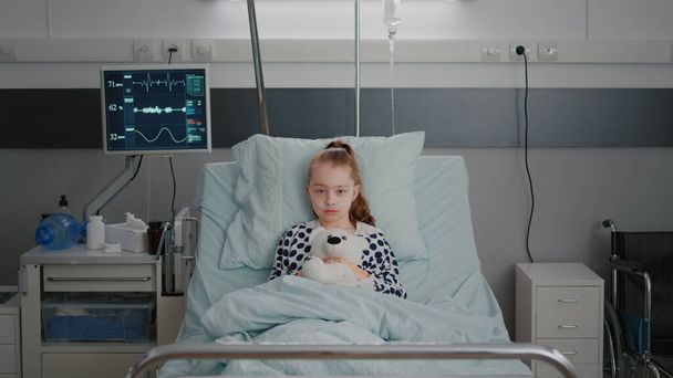 Porträt eines kranken Mädchens im Krankenhaus, das einen Teddybär hält, der während der ärztlichen Beratung im Bett liegt - Foto, Bild