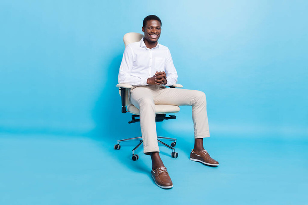 Photo of confident positive dark skin man wear white shirt smiling sitting chair isolated blue color background - Valokuva, kuva