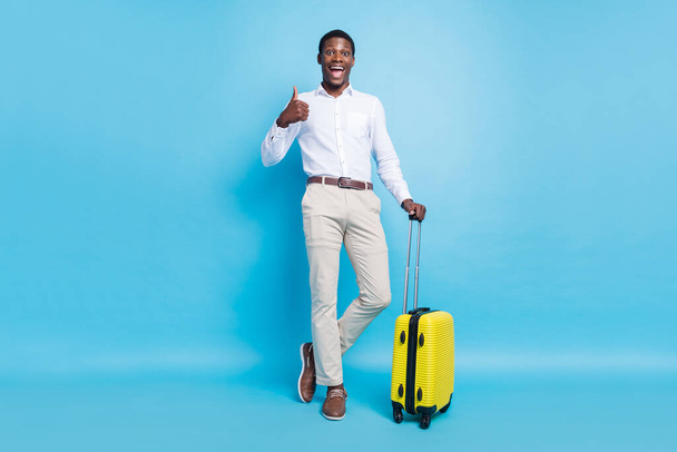 Photo of excited funny dark skin man wear white shirt smiling holding baggage showing thumb up isolated blue color background - Photo, Image