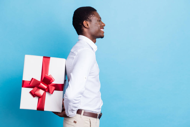 Photo of funny cute dark skin man wear white shirt smiling hiding big present box looking empty space isolated blue color background - Foto, Imagem