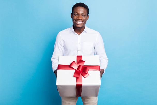 Photo of cute charming dark skin guy dressed formal shirt presenting you gift box smiling isolated blue color background - Fotografie, Obrázek