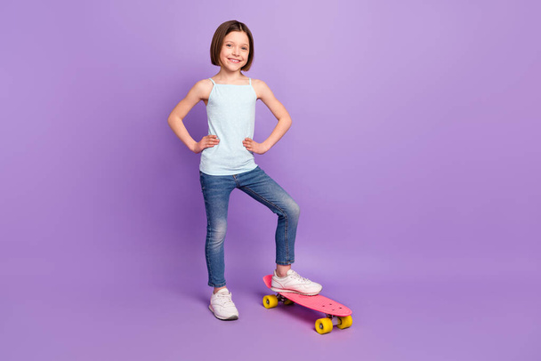 Full length photo of cheerful young nice girl hold hands waist stand longboard isolated on purple color background - Foto, imagen
