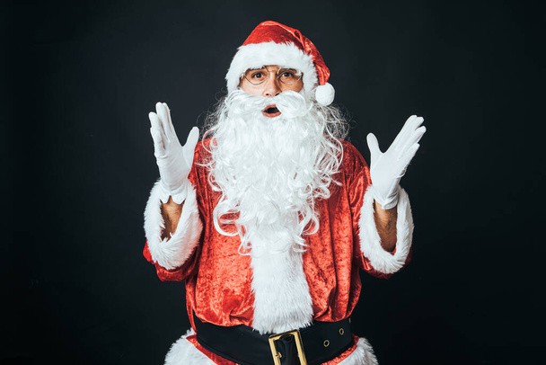 Человек, одетый как Санта Клаус, удивлен, с поднятыми руками, на черном фоне. Рождественская концепция, Санта-Клаус, подарки, праздник. - Фото, изображение