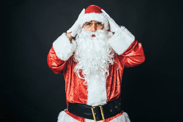Uomo vestito da Babbo Natale sorpreso con le mani sulla testa, su sfondo nero. Concetto di Natale, Babbo Natale, regali, celebrazione. - Foto, immagini