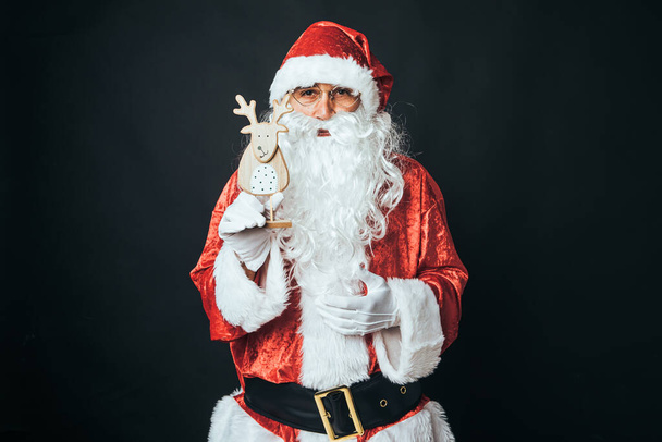 Άνδρας ντυμένος Άγιος Βασίλης κρατώντας τη φιγούρα ενός ξύλινου ταράνδου, σε μαύρο φόντο. Χριστούγεννα έννοια, Άγιος Βασίλης, δώρα, γιορτή. - Φωτογραφία, εικόνα
