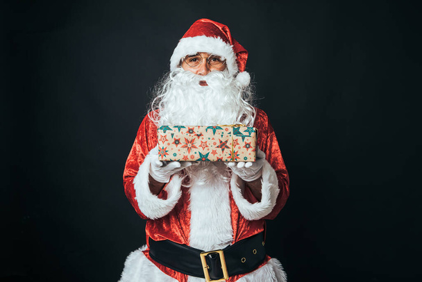 Άνδρας ντυμένος Άγιος Βασίλης κρατώντας ένα δέμα τυλιγμένο με χαρτί περιτυλίγματος, σε μαύρο φόντο. Χριστούγεννα έννοια, Άγιος Βασίλης, δώρα, γιορτή. - Φωτογραφία, εικόνα