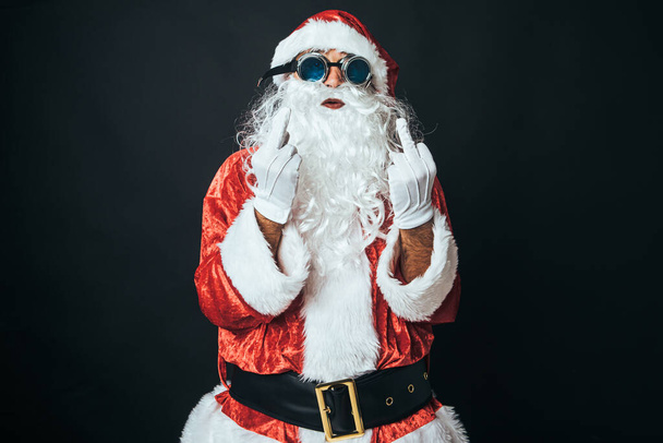 Man dressed as Santa Claus making a rude gesture, raise his fingers as a sign of 'fuck you', on black background. Christmas concept, Santa Claus, gifts, celebration. - Photo, Image