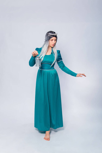 Full length portrait of a princess in a medieval, fantasy, turquoise dress with ash hair and a silver crown, posing with a lamp in her hands isolated on a white background. - Фото, изображение
