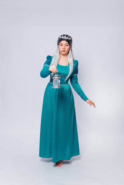 Full length portrait of a princess in a medieval, fantasy, turquoise dress with ash hair and a silver crown, posing with a lamp in her hands isolated on a white background. - Photo, image