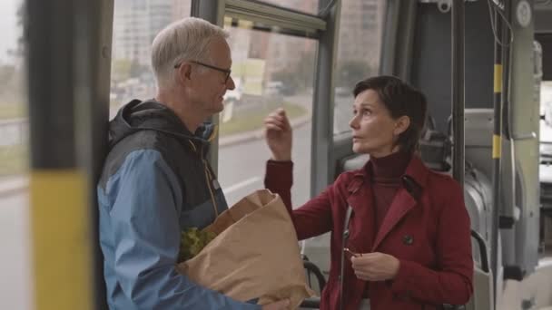 Meio de homem sênior segurando saco de papel com mantimentos e mulher caucasiana vestindo casaco vermelho em pé à janela no ônibus, tendo conversa - Filmagem, Vídeo