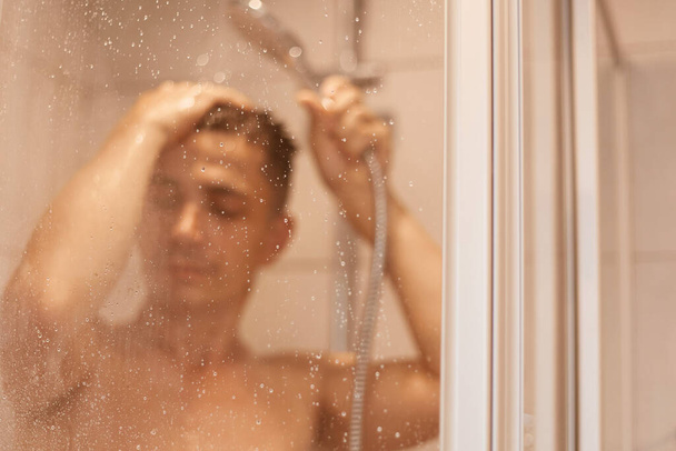 Indoor shot of young adult good looking and attractive man with wet body taking shower in bath, keeping eyes closed, washing hair, relaxed under hot water. - Photo, Image