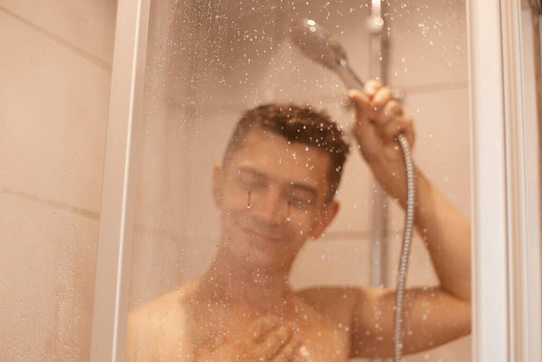 Happy smiling satisfied man posing naked in the shower behind glass with drops, taking shower in the morning in bathroom, refreshment after hard working day. - 写真・画像