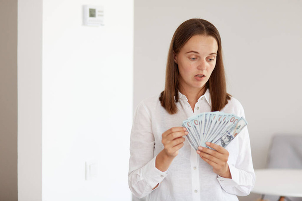 Portrait of astonished young adult beautiful girl wearing white casual style shirt, standing holding big sum of money, having surprised facial expression. - Photo, Image