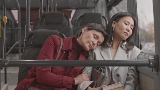 Waist-up of grey-haired Caucasian woman wearing red trench coat, sleeping on shoulder of young female Asian companion who also napping, then waking up and apologizing for it, sitting in bus - Footage, Video