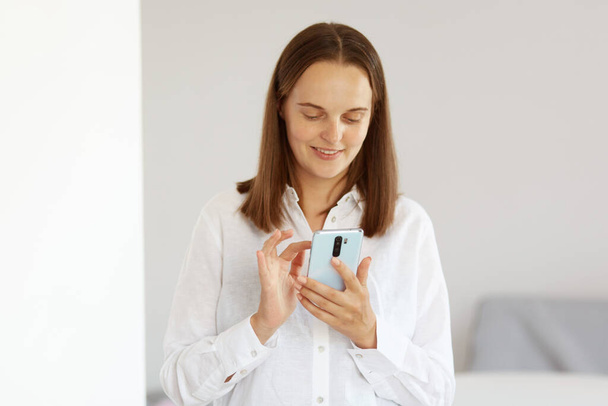 Indoor shot of smiling woman wearing white casual style shirt, holding cell phone in hands, looking at device screen, typing message or writing post, posing in light room at home. - Photo, Image