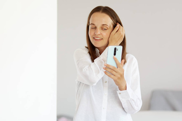 Portret atrakcyjnej szczęśliwej kobiety w białej, luźnej koszuli, trzymającej telefon komórkowy w rękach, robiącej selfie, patrzącej na wyświetlacz smartfona, pozującej w świetlicy w domu. - Zdjęcie, obraz