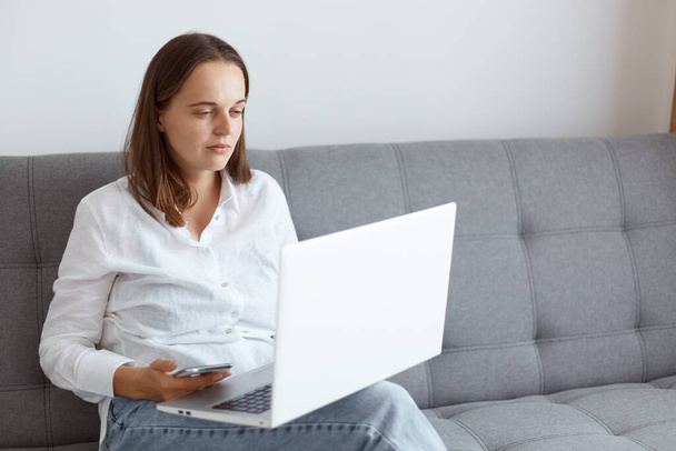Portrait dark haired female wearing white casual style shirt and jeans, sitting on sofa in living room, looking at laptop display, having problems with eyesight. - Foto, Imagen