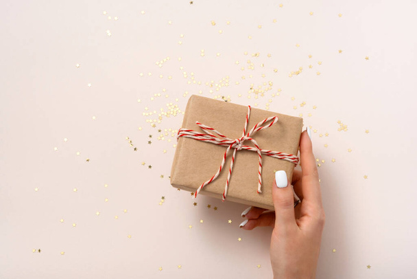 La mano femenina sostiene la caja de regalo en papel artesanal con cinta blanca-roja de Año Nuevo y estrellas de la confitería de oro en un espacio de copia de fondo beige, disposición plana. Navidad, boda - Foto, imagen