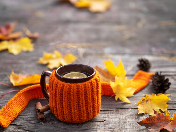 Чашка горячего чая с лимоном и корицей в вязаном оранжевом свитере или куртке. На деревянном столе в лесу, сезон золотой осени. Желтые кленовые листья - Фото, изображение