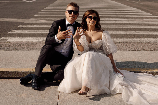 молодята в коханні, наречений і наречений у весільній сукні сидять на пішохідному переході на тлі міського пейзажу і показують обручки на пальцях, концепція весілля сонцезахисні окуляри на очах
.  - Фото, зображення