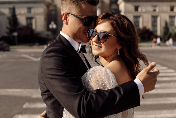 Foto van jong stel, bruid en bruidegom in zonnebril, als speciale agenten in trouwjurk. er is stadslandschap op de achtergrond. bruiloften. gelukkige momenten - Foto, afbeelding