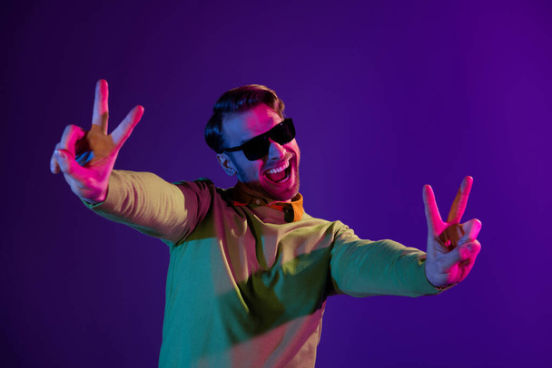 Photo portrait of smiling man wearing sunglass showing peace v-sign smiling isolated on dark purple color background - Фото, зображення