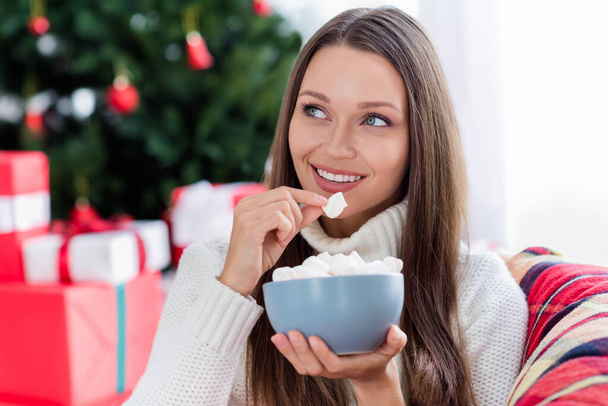 Foto de linda mujer joven vestida suéter de punto comiendo dulces sonriendo en el interior habitación casa - Foto, Imagen