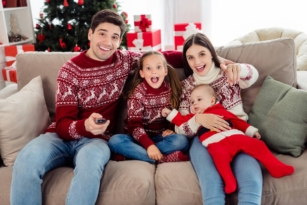 Photo of impressed cute couple two kids dressed new year pullovers sitting couch watching tv pointing finger you indoors house home room - Foto, Imagen