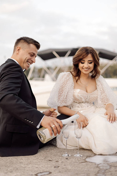young couple, bride and groom in wedding dresses are sitting on concrete bridge with glasses of wine and champagne. spend time together, happy. - Foto, Imagen