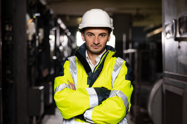 Young factory engineer or worker wearing safety vest and hard hat crossing arms at electrical control room. Industry and engineering concept - Фото, зображення