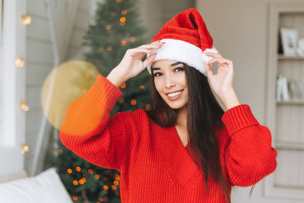 Young beautiful asian woman with dark long hair in cozy red knitted sweater and santa hat having fun in room with Christmas tree - Photo, image