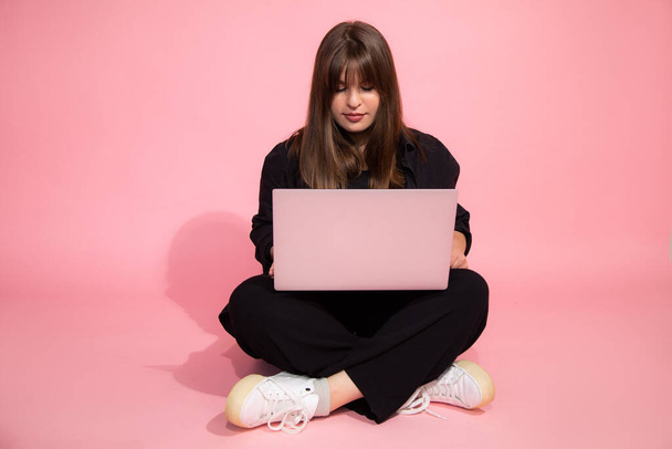 学生茶色の髪はピンクの背景に座ってノートパソコンを保持カジュアル服で魅力的な女の子を驚かせた。オンライン教育の概念。スタジオショット. - 写真・画像