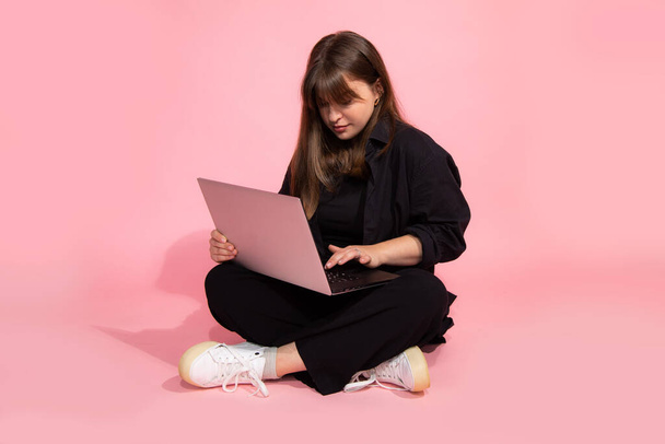  Diák barna hajú lány alkalmi ruhák gazdaság Laptop számítógép ül a rózsaszín háttér. Online oktatási koncepció. Stúdió lövés.  - Fotó, kép