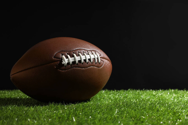 American football ball on green grass against black background. Space for text - Φωτογραφία, εικόνα