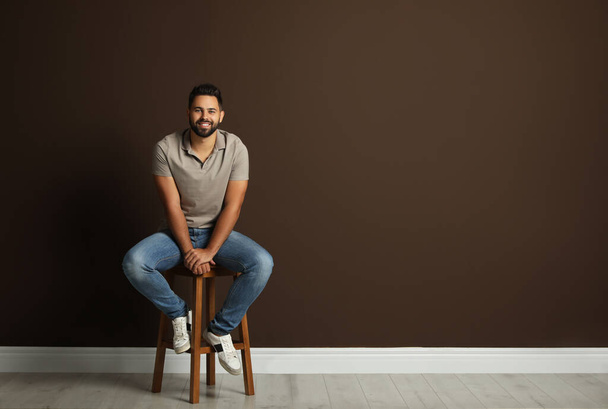 Handsome young man sitting on stool near brown wall. Space for text - Foto, Bild