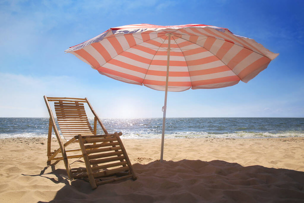 Deck chair near red and white striped beach umbrella on sandy seashore - Photo, image