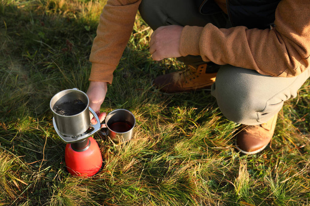 Man making hot drink with portable gas burner on green grass, closeup. Camping season - Foto, Bild