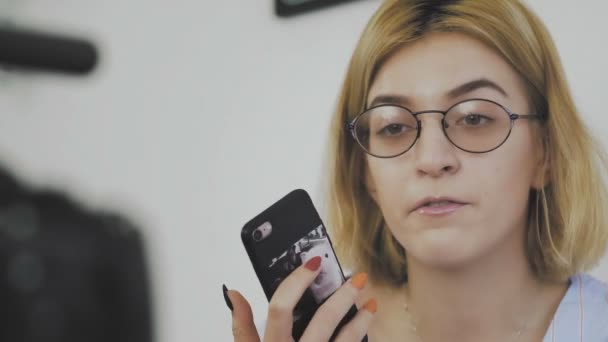 Gros plan d'une jeune femme avec des lunettes assis avec un téléphone dans les mains, souriant et parlant à la caméra - Séquence, vidéo