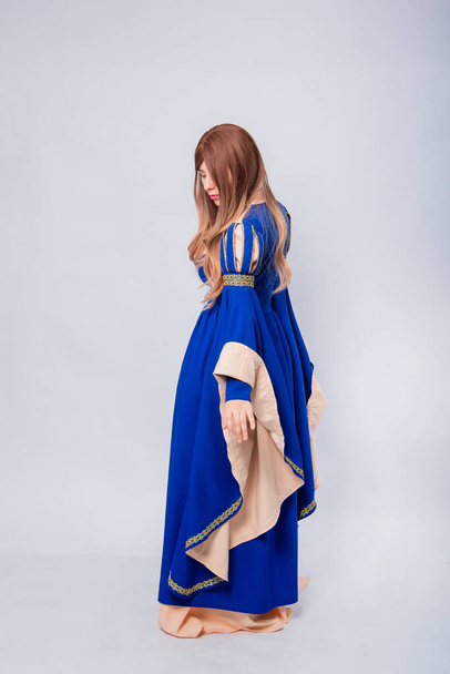 A full-length portrait of an attractive woman with long hair standing back in a medieval, fantastic, blue and beige dress with long flared sleeves, posing isolated on a white background. - Foto, imagen