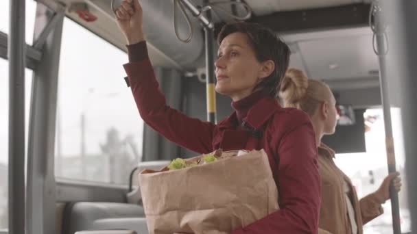 Bajo ángulo de mujer caucásica de pelo corto con gabardina roja, bolsa de papel con comestibles, autobús - Imágenes, Vídeo